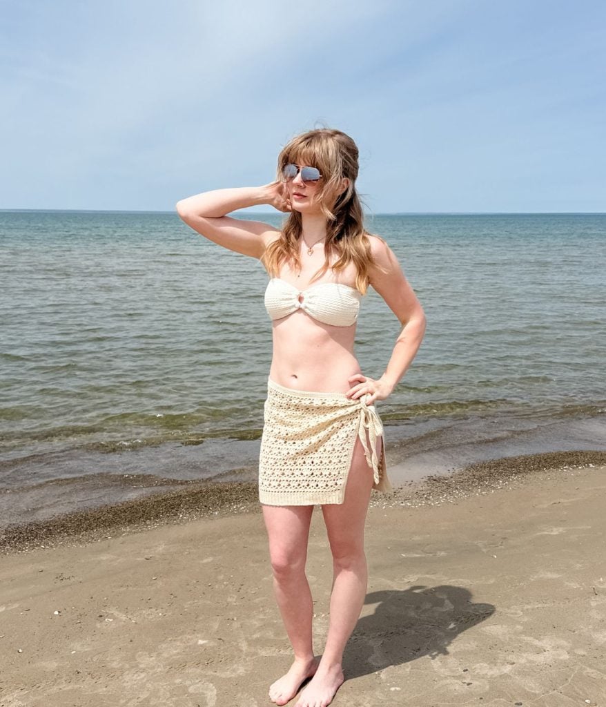 A woman in a cream bikini top and Caribbean Seashell skirt crochet pattern stands barefoot on a sandy beach near the water, with one hand on her hip and the other near her head.