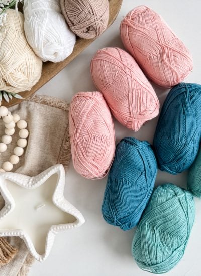 Yarn skeins in shades of pink, blue, and green are arranged on a white surface beside a star-shaped candle, beige tassels, wooden beads, and a wooden tray with neutral yarn and greenery from Mjs Off The Hook Designs.