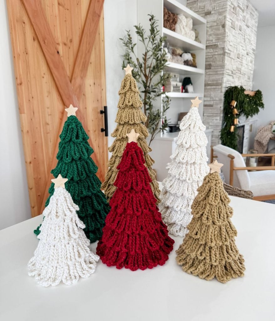 Six bulky crochet Christmas tree decorations in green, red, and white yarn with star toppers are displayed on a white table in a modern living room setting. Perfect for adding festive charm with this unique crochet pattern.