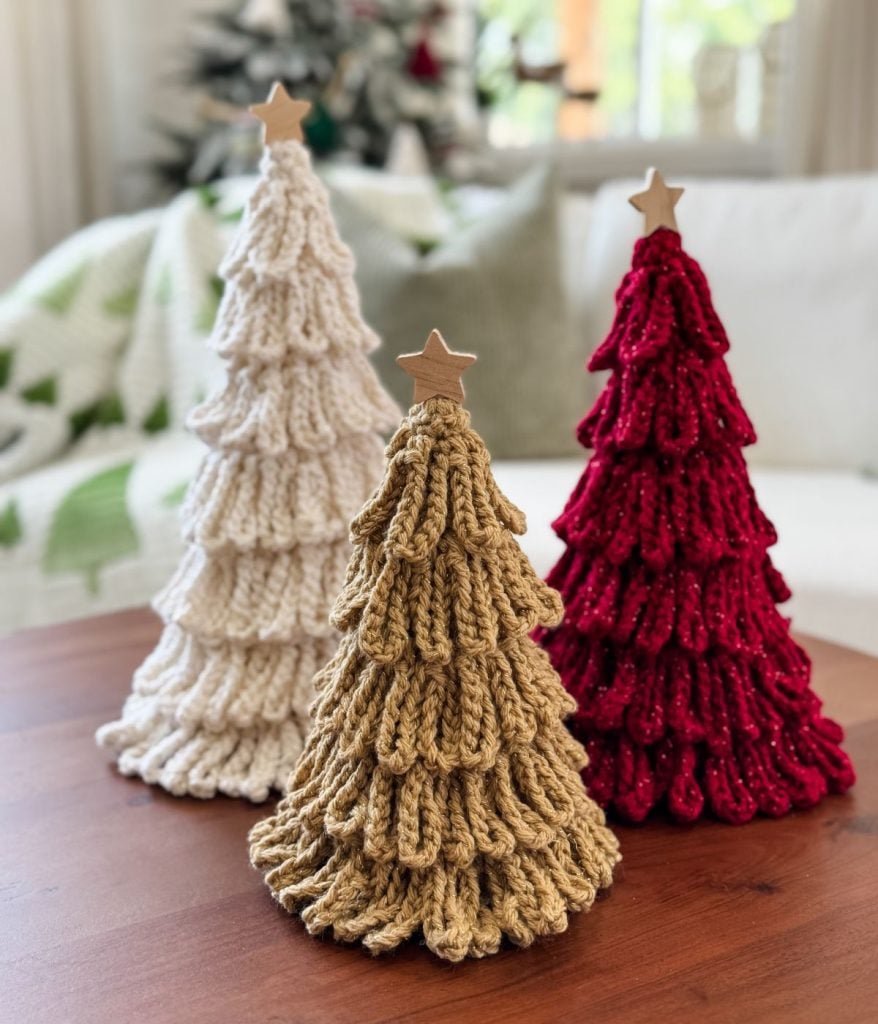 Three crocheted Christmas trees—white, gold, and red—made with a bulky crochet pattern and topped with wooden stars, are displayed on a wooden table with a cozy couch and blanket in the background.