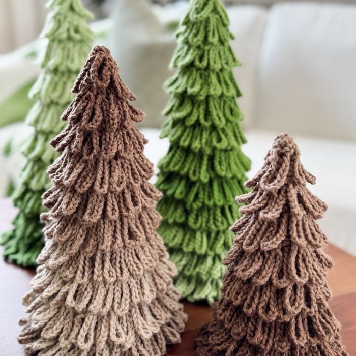 Four crochet trees in varying shades of green and brown yarn are displayed on a wooden surface, with a blurred sofa in the background.