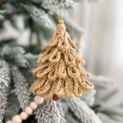 A Loopy Tree crocheted Christmas ornament hangs on a snow-dusted pine branch, with a string of wooden beads visible in the background.