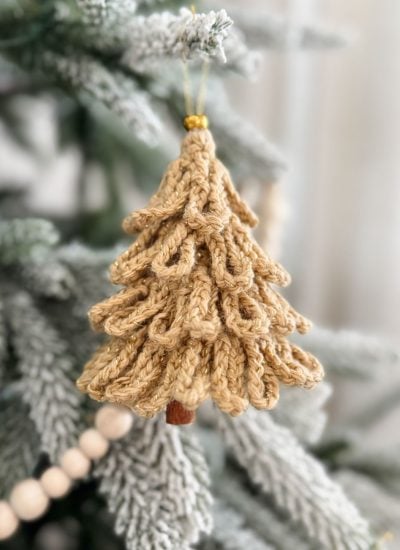 A Loopy Tree crocheted Christmas ornament hangs on a snow-dusted pine branch, with a string of wooden beads visible in the background.