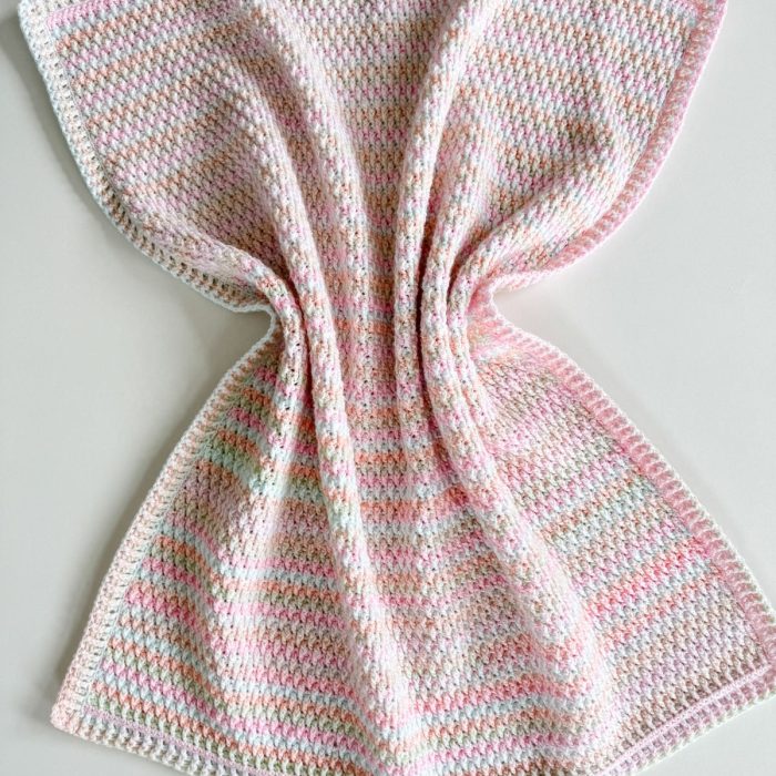 A crocheted Alpine Stitch Blanket with a textured pattern in pastel shades of pink, white, and light green, laid flat on a light surface.
