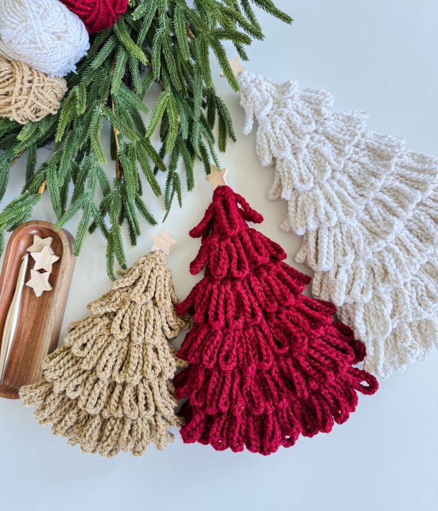 Three Loopy Crochet Trees in beige, red, and white are arranged next to evergreen branches, with a small tray holding two star-shaped ornaments nearby.
