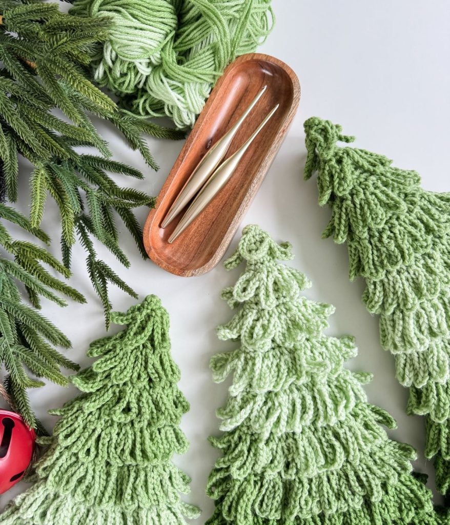 Crochet Trees in green yarn are arranged near a wooden tray holding two crochet hooks, a ball of green yarn, and artificial evergreen branches.