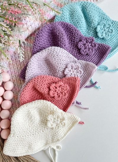 Five crocheted baby bonnets in pastel colors—blue, purple, lilac, pink, and white—each with a flower detail, are laid out like a baby sunhat collection on a white surface next to decorative beads and flowers.