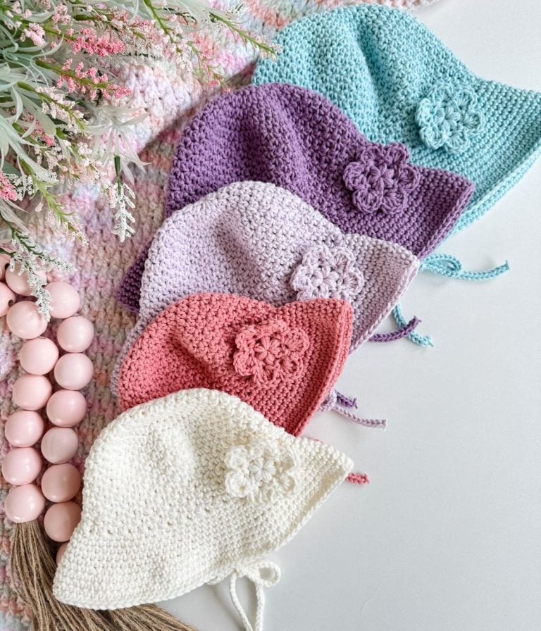 Five crocheted baby bonnets in pastel colors—blue, purple, lilac, pink, and white—each with a flower detail, are laid out like a baby sunhat collection on a white surface next to decorative beads and flowers.