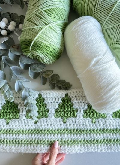 A hand holds a crocheted piece with green and white yarn, featuring a row of green tree patterns—evoking a Winter Forest. Two balls of yarn and some eucalyptus leaves are placed nearby.