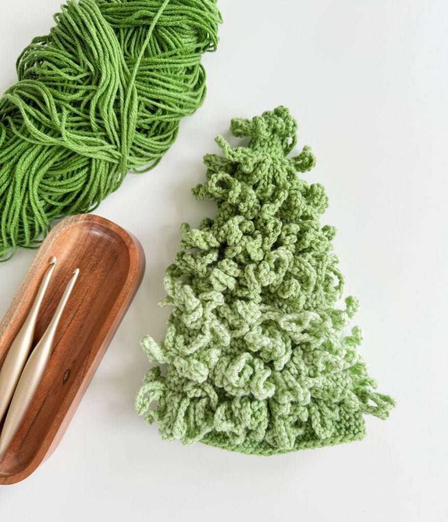 A green, textured crocheted piece shaped like a tree sits next to a skein of green yarn and two crochet hooks in a wooden tray on a white surface, creating the perfect scene for crafting beautiful Crochet Trees.