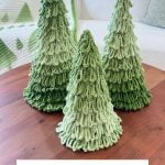 Three green Loopy Tree crochet trees with ombre patterns are displayed on a wooden table, with a green and white tree-patterned blanket in the background.
