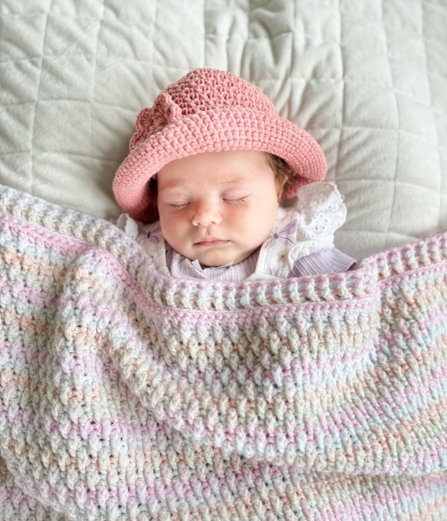 A baby wearing a pink knit baby sunhat sleeps under a pastel-colored knit blanket on a quilted white surface.