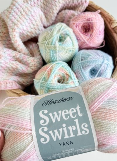 A hand holds a skein of Herrschners Sweet Swirls yarn in front of a basket filled with pastel-colored yarn balls and a partially completed Alpine Stitch Blanket crochet project.