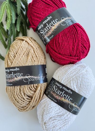 Three skeins of Mary Maxim Starlette Sparkle yarn in red, gold, and white are arranged next to pine branches—perfect for creating quick crochet trees or festive bulky crochet patterns.