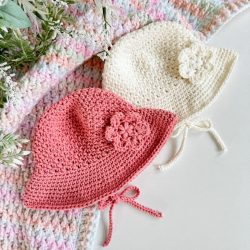 Two crocheted baby hats, one pink and one cream, each designed as a charming baby sunhat with a flower accent and ties, are displayed on a pastel crocheted blanket with some greenery.