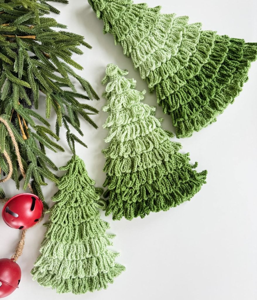 Three green, loop-stitched crochet trees are arranged on a white surface next to pine branches and two red jingle bells. These charming crochet Christmas trees add a festive touch to your holiday decor.