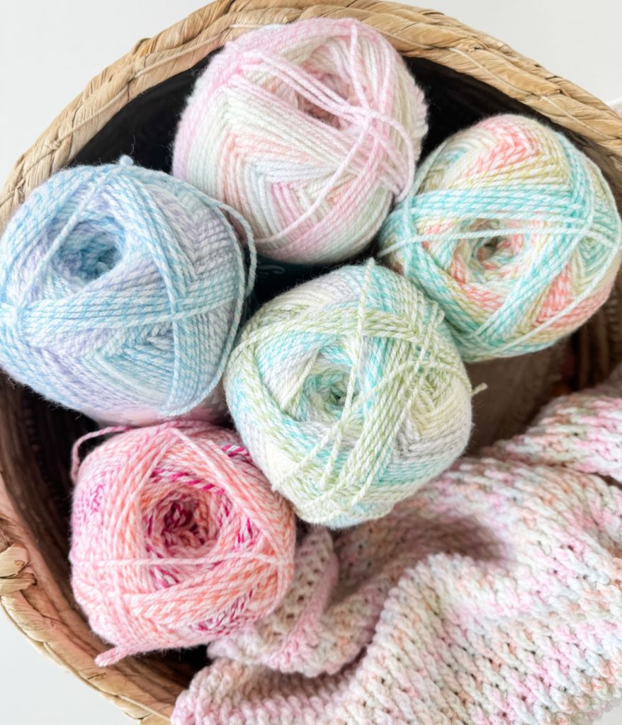 Five pastel-colored yarn balls in a woven basket sit next to a partially knitted Alpine Stitch Blanket, showcasing cozy creativity in progress.