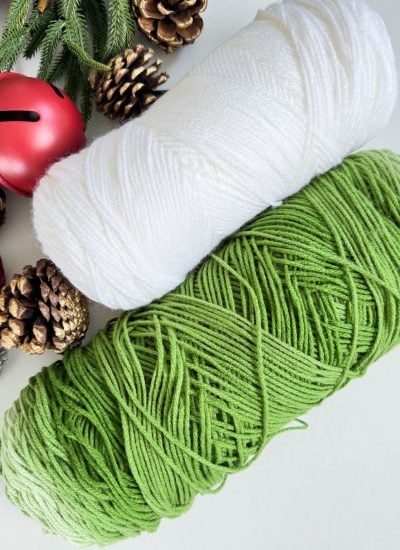 Two skeins of yarn, one white and one green, are placed on a white surface with pinecones, fir branches, red jingle bells, and finished Crochet Trees for a festive touch.