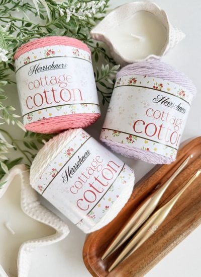 Three skeins of Herrschners Cottage Cotton yarn in pink, white, and lavender rest on a white surface with candles, greenery, two gold crochet hooks—perfect colors for making a sweet baby sunhat.