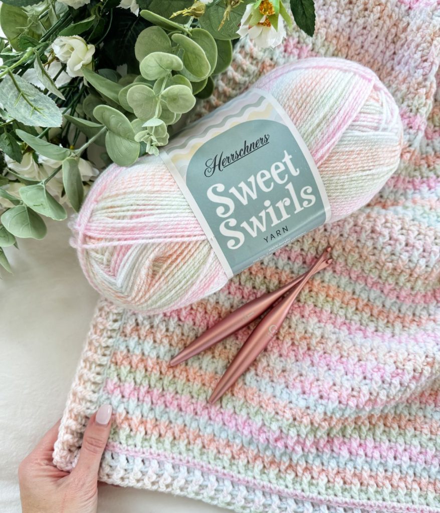 A skein of pastel Sweet Swirls yarn, an Alpine Stitch Blanket in progress, two rose gold crochet hooks, and a bouquet of green and white foliage.