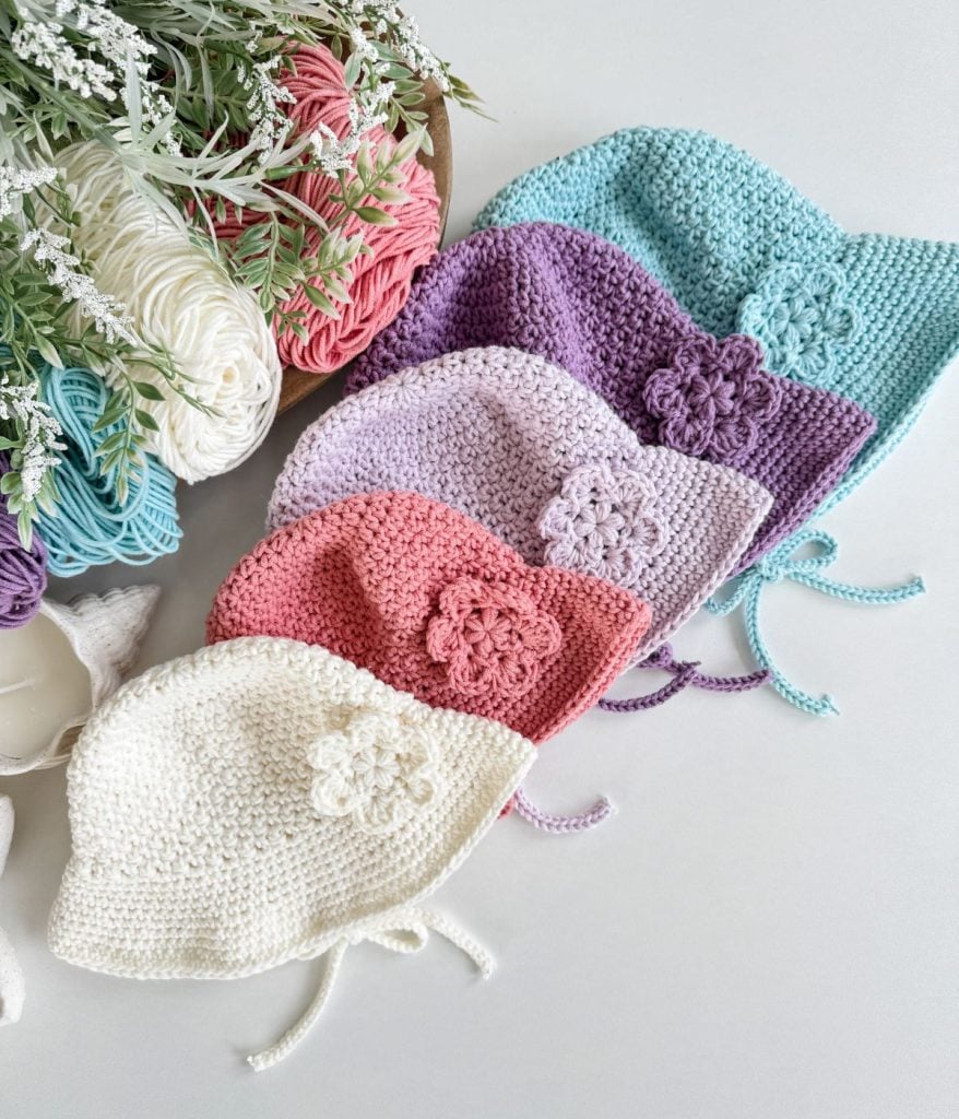 Five crocheted baby bonnets, including a sweet baby sunhat in white, pink, lavender, purple, and blue—each adorned with a flower—are displayed beside yarn and faux greenery.
