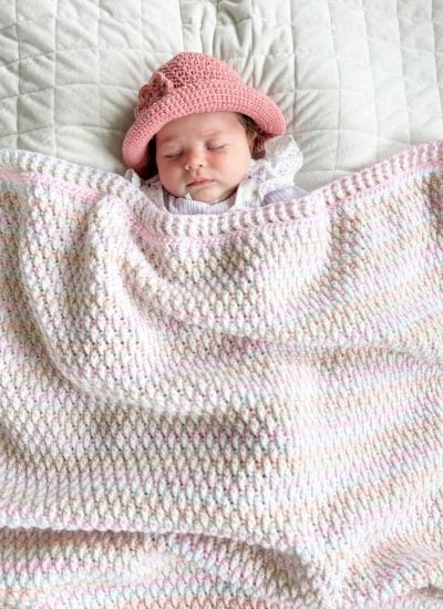 A baby wearing a pink crocheted hat sleeps on a quilted surface, snuggled under a pastel Alpine Stitch Blanket.