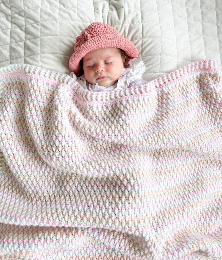 A baby wearing a pink crocheted hat sleeps on a quilted surface, snuggled under a pastel Alpine Stitch Blanket.