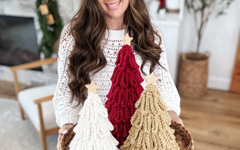 A woman holds a woven tray with three handmade loopy crochet trees in white, red, and beige, standing in a cozy, decorated living room.