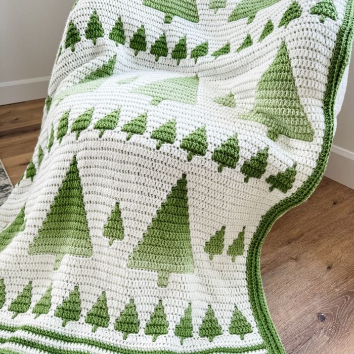 A crocheted Mosaic Blanket with green and white stripes, featuring rows of large and small green trees reminiscent of a Winter Forest, is draped over a chair on a wooden floor.