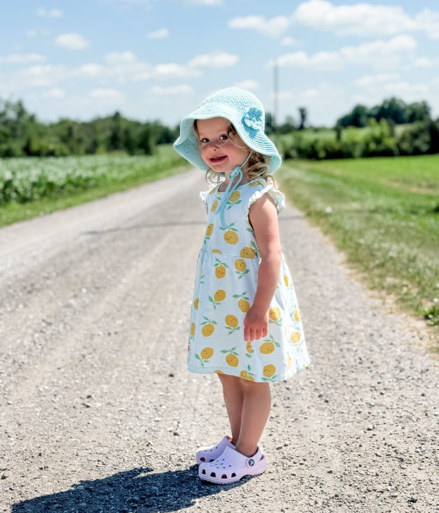 A young child in a lemon-print dress and baby sunhat stands on a gravel road outdoors, with grass and trees in the background under a blue sky.