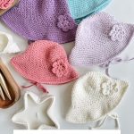 Five crocheted baby sunhats in pastel colors are arranged on a white surface, each featuring a decorative flower and ties. Text below reads: "Baby Sand Sunhat Pattern, Crochet Pattern & Video.
