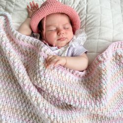 A baby wearing a pink hat sleeps on a white quilt, partially covered with a pastel-colored Alpine Stitch Blanket.