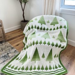 A Mosaic Blanket with green tree patterns evokes a Winter Forest, draped over a chair in a bright room with wooden floors and a potted plant.