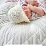 A sleeping baby wearing a crocheted white baby sunhat lies on a quilted blanket with a pink and white crochet blanket partially covering them. Text reads "Baby Sand Sunhat Pattern.