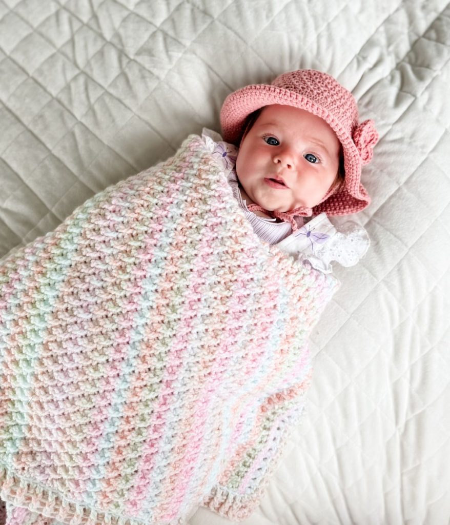 A baby wearing a pink hat is wrapped in a pastel Alpine Stitch Blanket, lying on a quilted white surface.