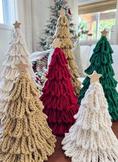 Five loopy crochet trees in beige, red, white, and green with wooden star toppers are displayed on a table, bringing handmade charm to your holiday décor with each unique bulky crochet tree, set against a festive tree in the background.