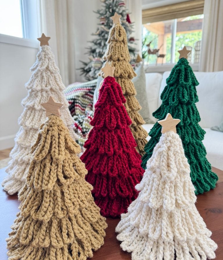 Five loopy crochet trees in beige, red, white, and green with wooden star toppers are displayed on a table, bringing handmade charm to your holiday décor with each unique bulky crochet tree, set against a festive tree in the background.
