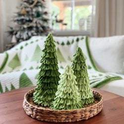 Three crochet trees in festive green sit in a woven basket on a wooden table, with a holiday-themed blanket and Christmas tree in the background.
