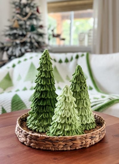 Three crochet trees in festive green sit in a woven basket on a wooden table, with a holiday-themed blanket and Christmas tree in the background.