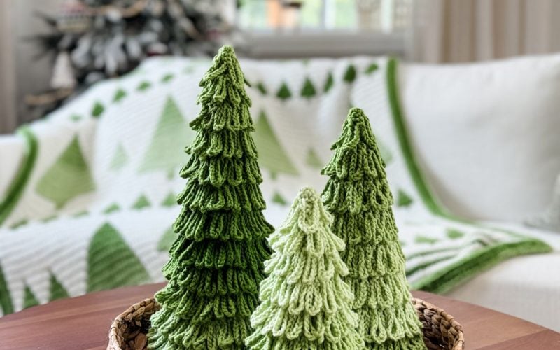 Three crochet trees in festive green sit in a woven basket on a wooden table, with a holiday-themed blanket and Christmas tree in the background.