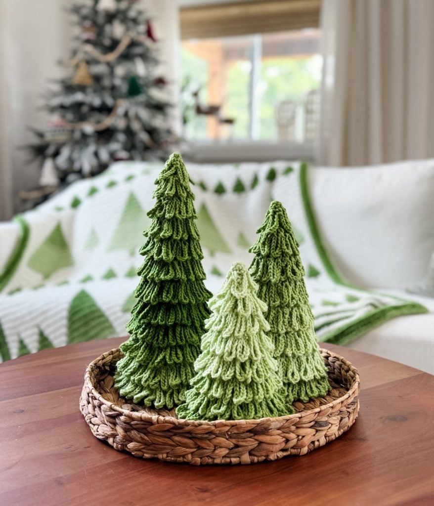 Three crochet trees in festive green sit in a woven basket on a wooden table, with a holiday-themed blanket and Christmas tree in the background.