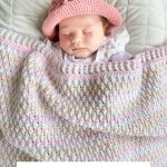 Baby wearing a pink crocheted baby sunhat sleeps under a pastel crocheted blanket on a light quilt. Text below reads: “BABY SAND SUNHAT PATTERN Crochet Pattern & Video.”.