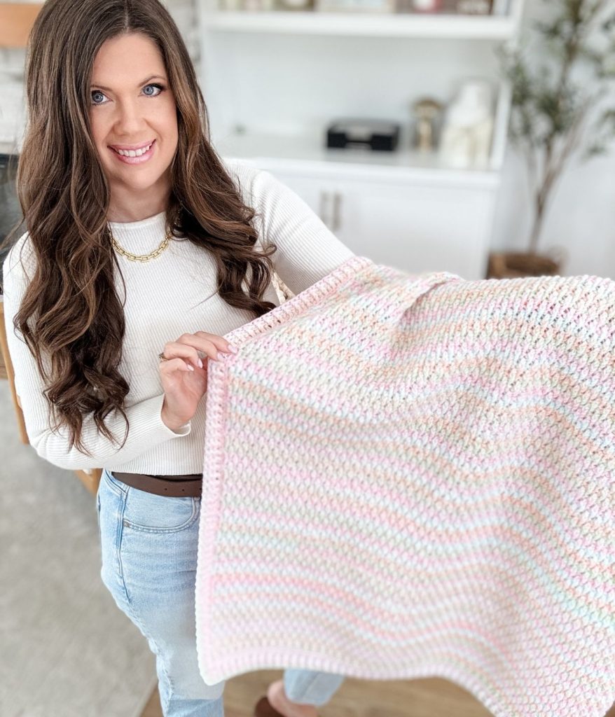 A woman with long brown hair and a white shirt holds up an Alpine Stitch Blanket in soft pink and white tones in a bright room.