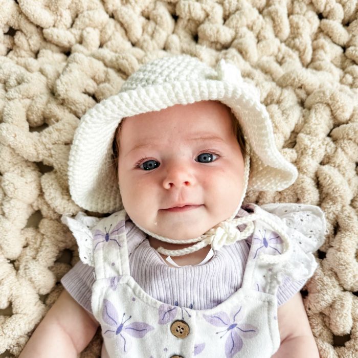 A baby wearing a white knitted baby sunhat and a floral-patterned outfit lies on a textured cream-colored blanket, looking at the camera.