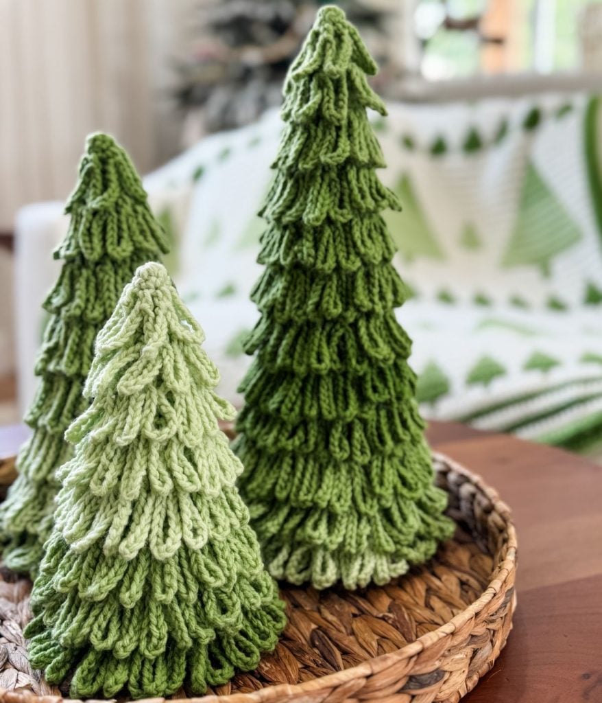 Three green crochet trees sit as Christmas decorations on a woven tray, with a crocheted blanket in the background.