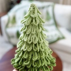 A hand holds a knitted green Christmas tree with layered branches; Crochet Trees and a blanket with tree patterns adorn the cozy couch in the background.