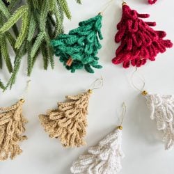 Loopy Tree crocheted Christmas ornaments in green, red, beige, and white are displayed next to artificial pine branches on a white surface.