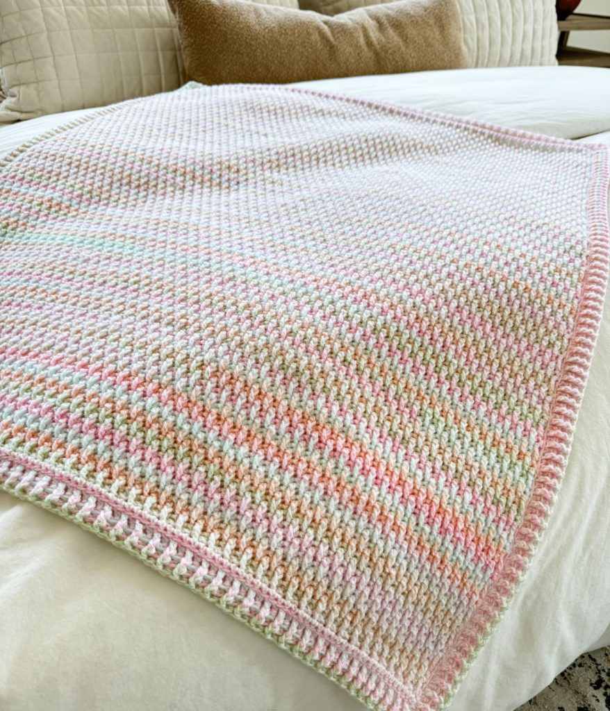 A pastel-colored Alpine Stitch Blanket with a textured pattern is spread neatly on a white bed, complemented by cream and tan pillows in the background.