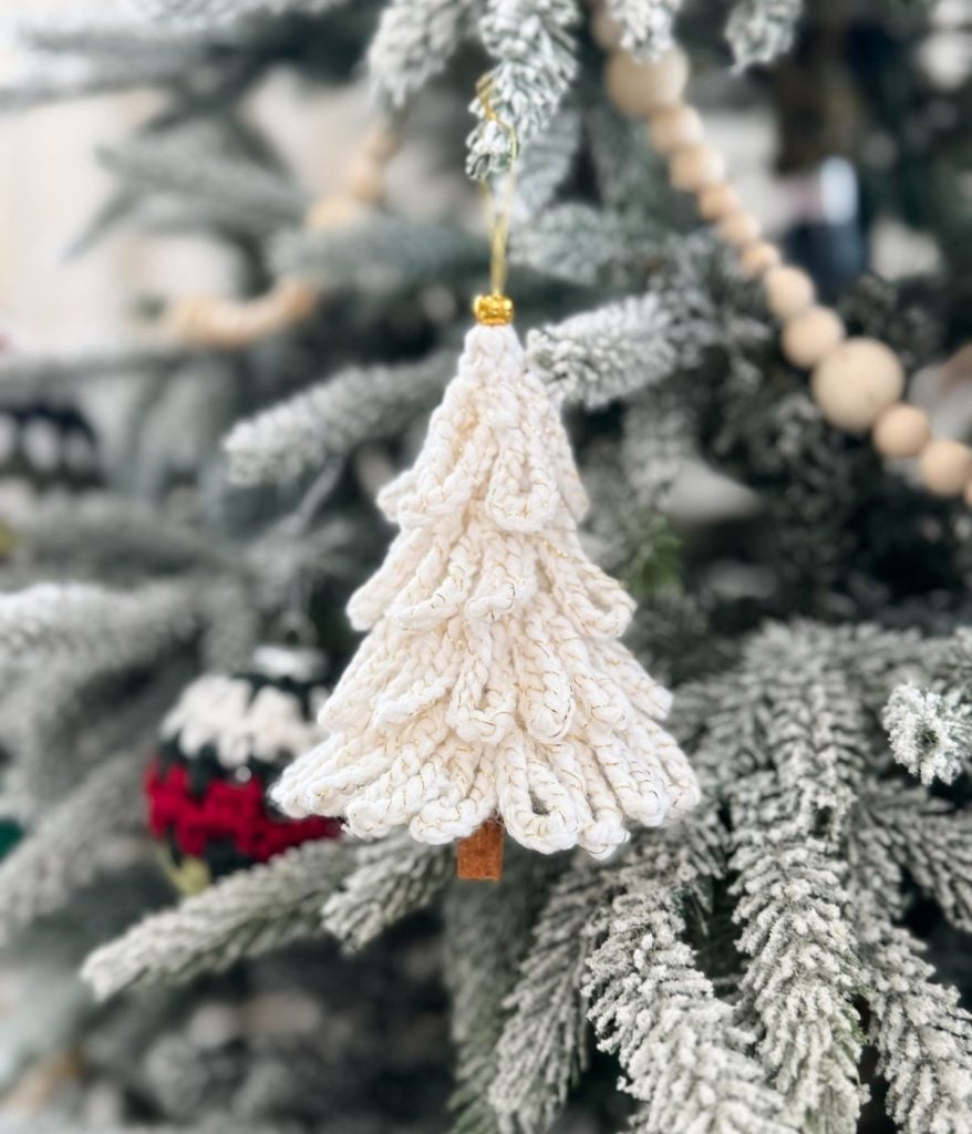 A white crocheted Loopy Tree Christmas ornament hangs on a snow-dusted evergreen branch, with wooden beads and other decorations in the background.