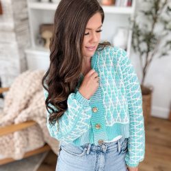 Woman with long brown hair wearing a seaglass mosaic-patterned cardigan and light blue jeans stands indoors in a cozy, well-decorated living space.